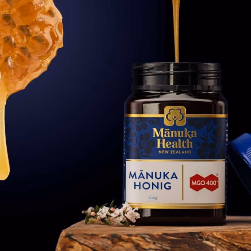 Mānuka Health Honigglas (MGO 400+) auf einem Holzpodest mit blauem Hintergrund und herabtropfendem Honig — Sinnbild für das Premium-Naturprodukt aus Neuseeland